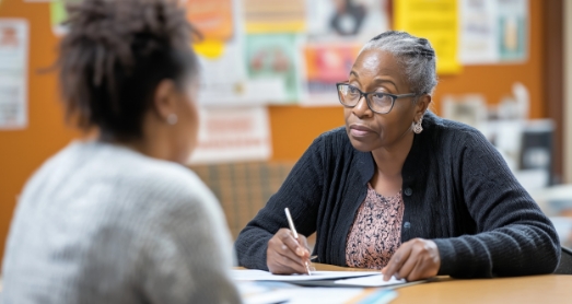 Two women sit across from each other at a table in an office, engaged in conversation. One woman writes on a notepad while listening attentively to the other. Bulletin boards with various papers are visible in the background.
